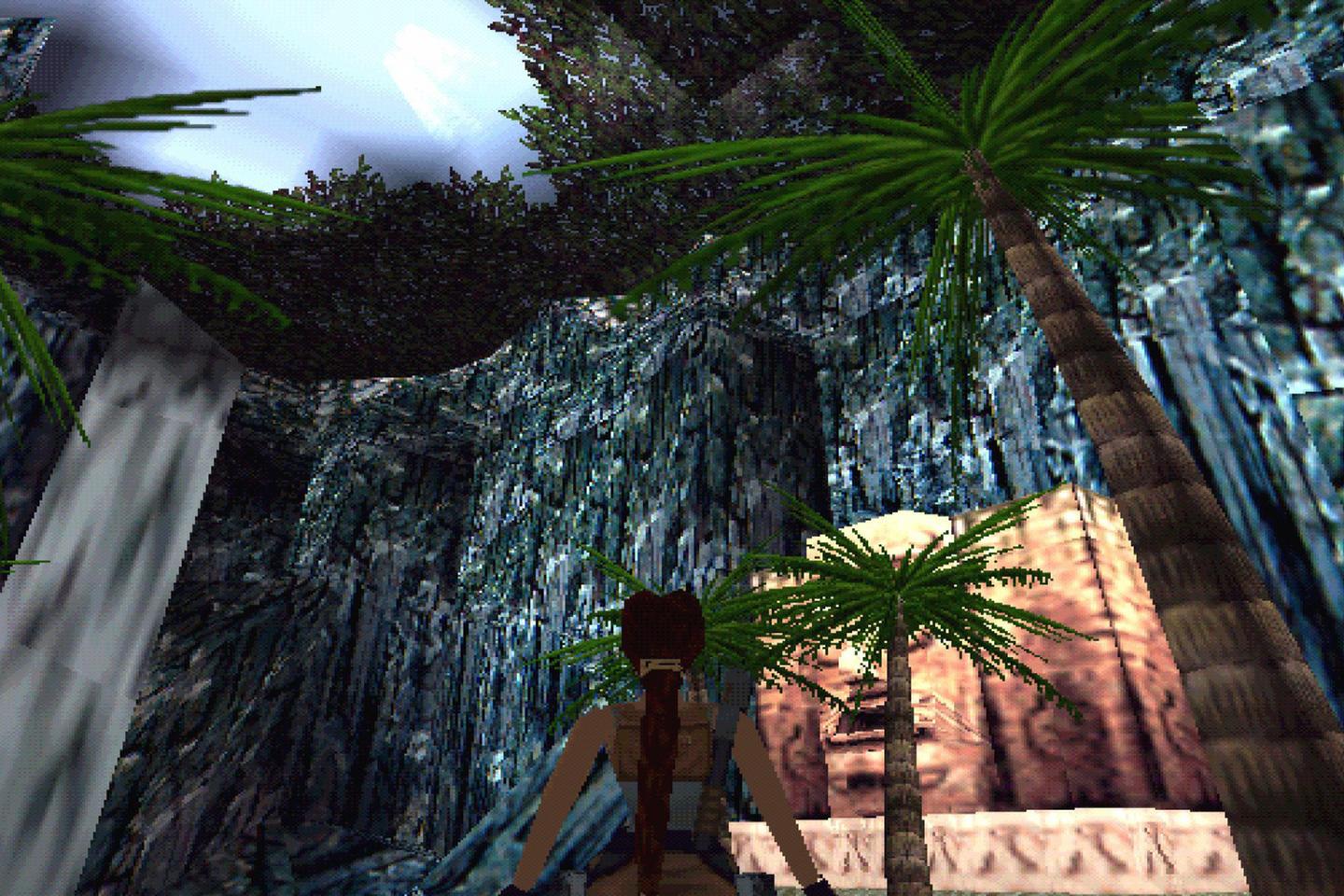 Lara with a ponytail stands near a palm tree, looking at a stone structure amidst towering rock walls under a sliver of sky.