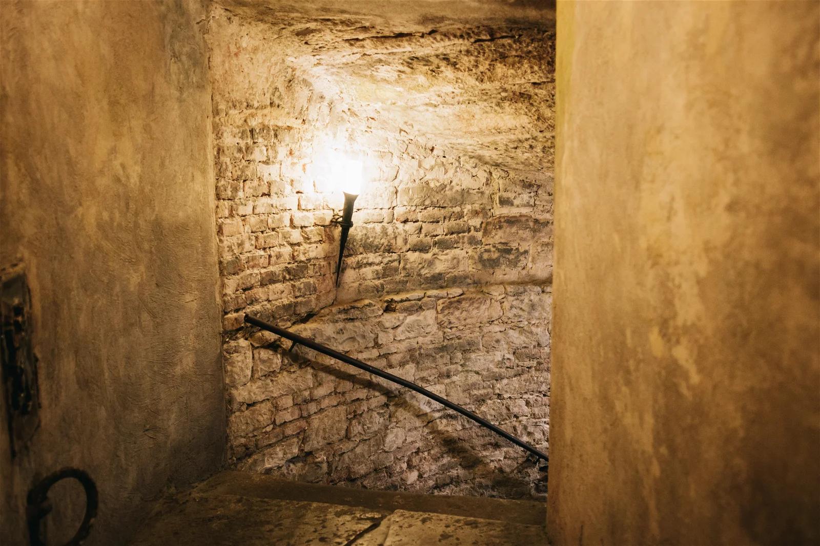 A photograph of a narrow, stone stairwell illuminated by a single wall lamp, evoking the feel of an ancient building or castle.