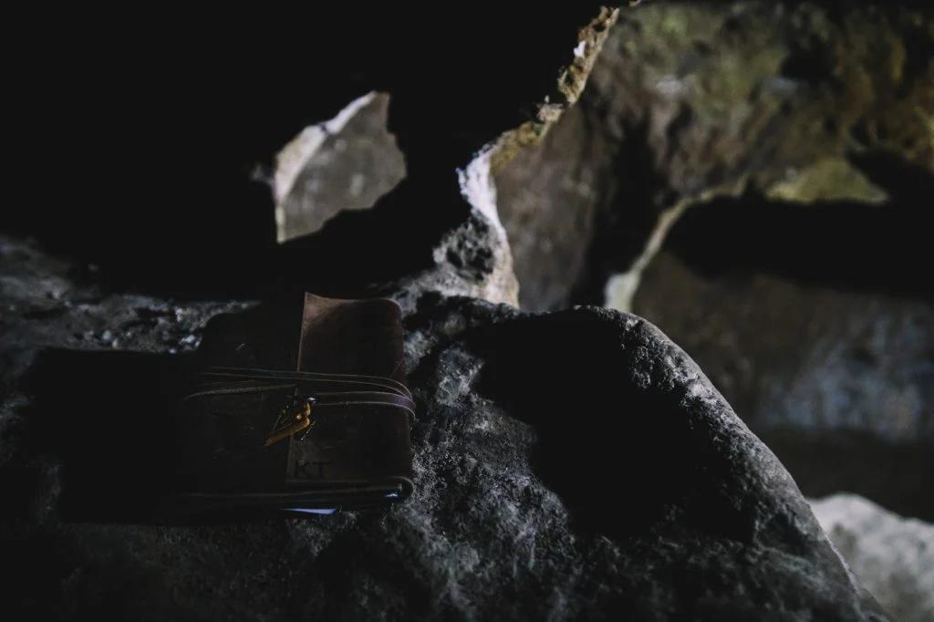 A close-up photo of a rugged leather satchel resting on a rocky surface in a dimly lit cave setting.