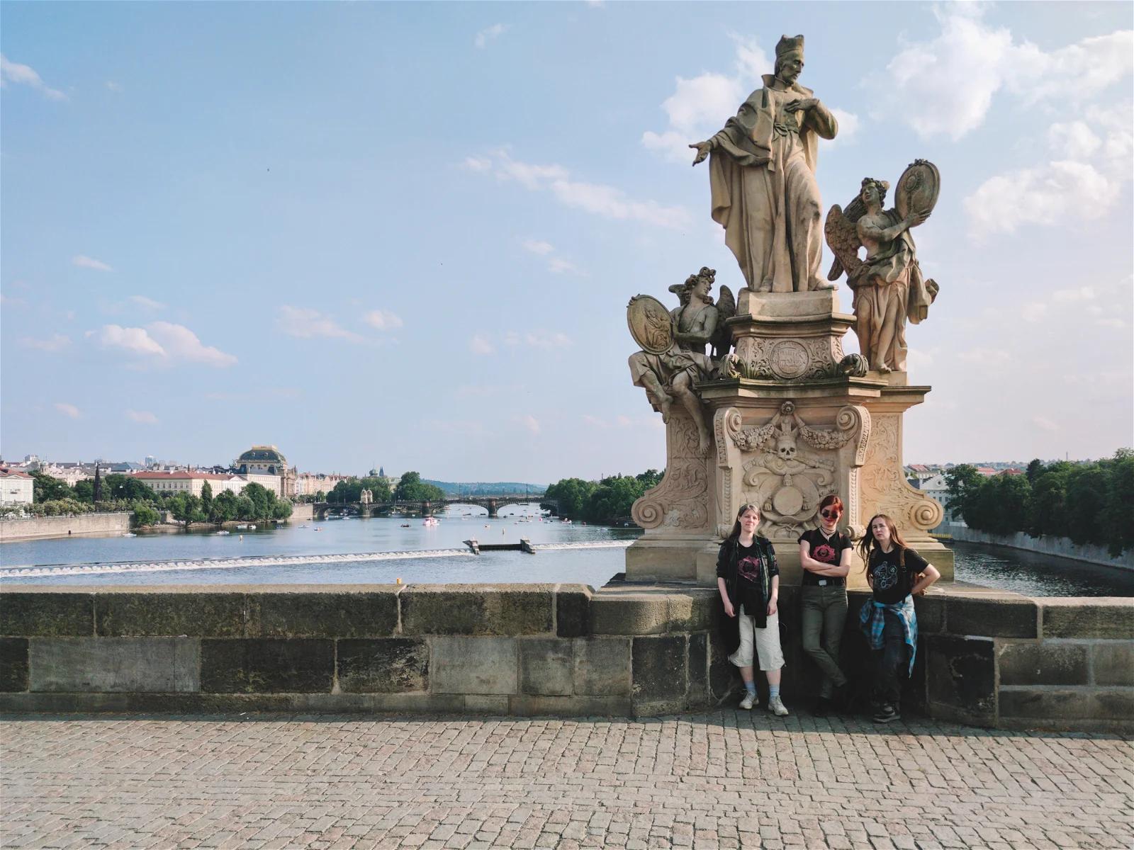 A photograph of people posing with a statue on a bridge, likely a tourist spot, with a river and cityscape in the background.