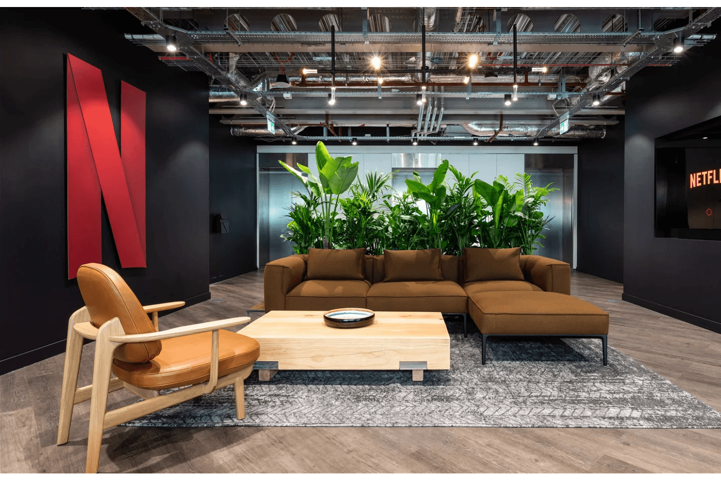 A modern office lounge with a large Netflix logo on the wall, featuring contemporary furniture and a wall of green plants under exposed ceiling infrastructure.