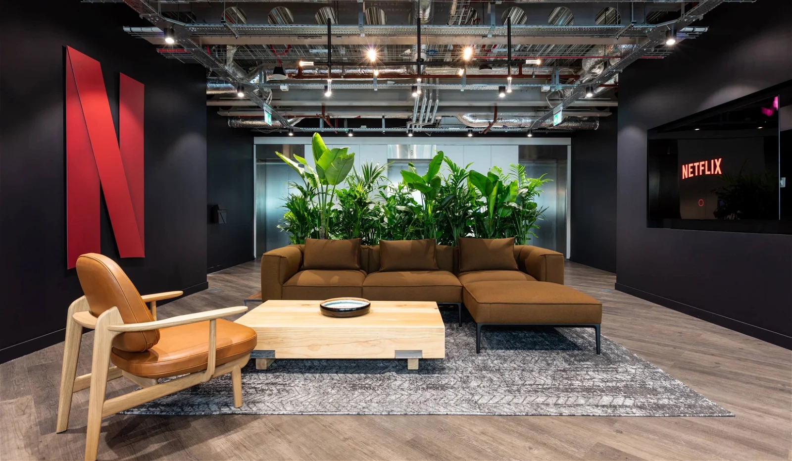 A modern office lounge with a large Netflix logo on the wall, featuring contemporary furniture and a wall of green plants under exposed ceiling infrastructure.