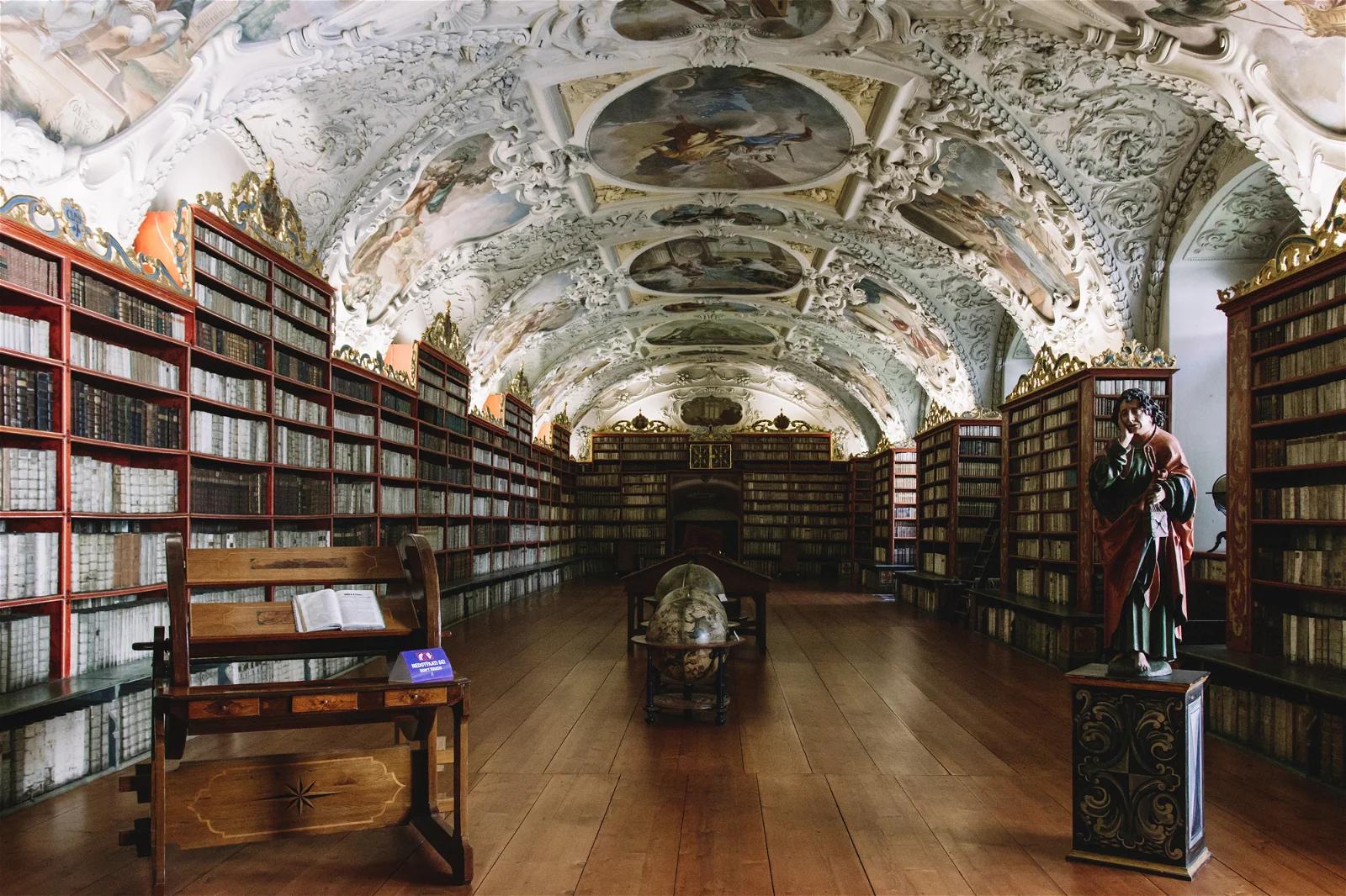 A grand, ornate library with a beautifully frescoed ceiling, filled with rows of wooden bookshelves housing numerous books, giving it a renaissance-era ambiance.
