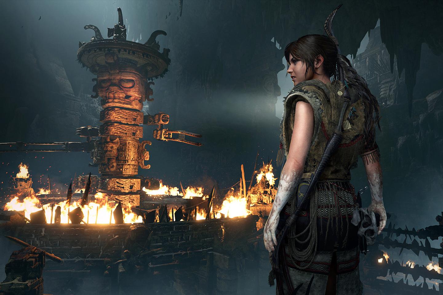 A moody in-game image from a Tomb Raider game capturing Lara Croft in tribal gear, gazing at a distant ritual site with lit torches and an ornate tower, set in an atmospheric cave.
