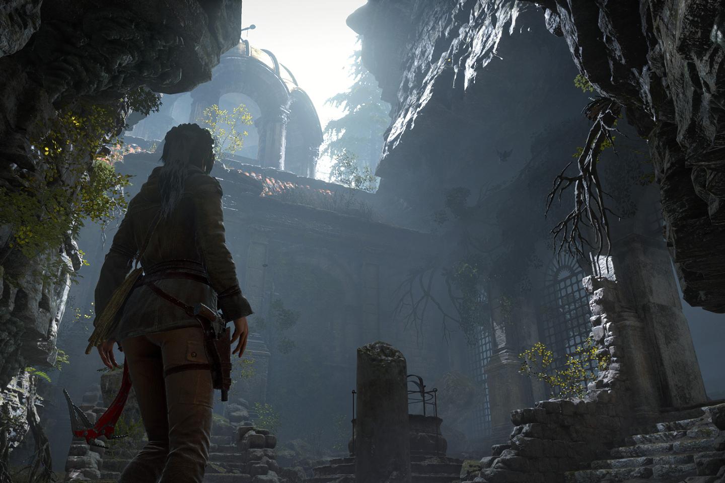 In-game screenshot of Lara Croft in Rise of the Tomb Raider, standing at the entrance of an ancient, sunlit temple surrounded by ruins and overgrown vegetation.