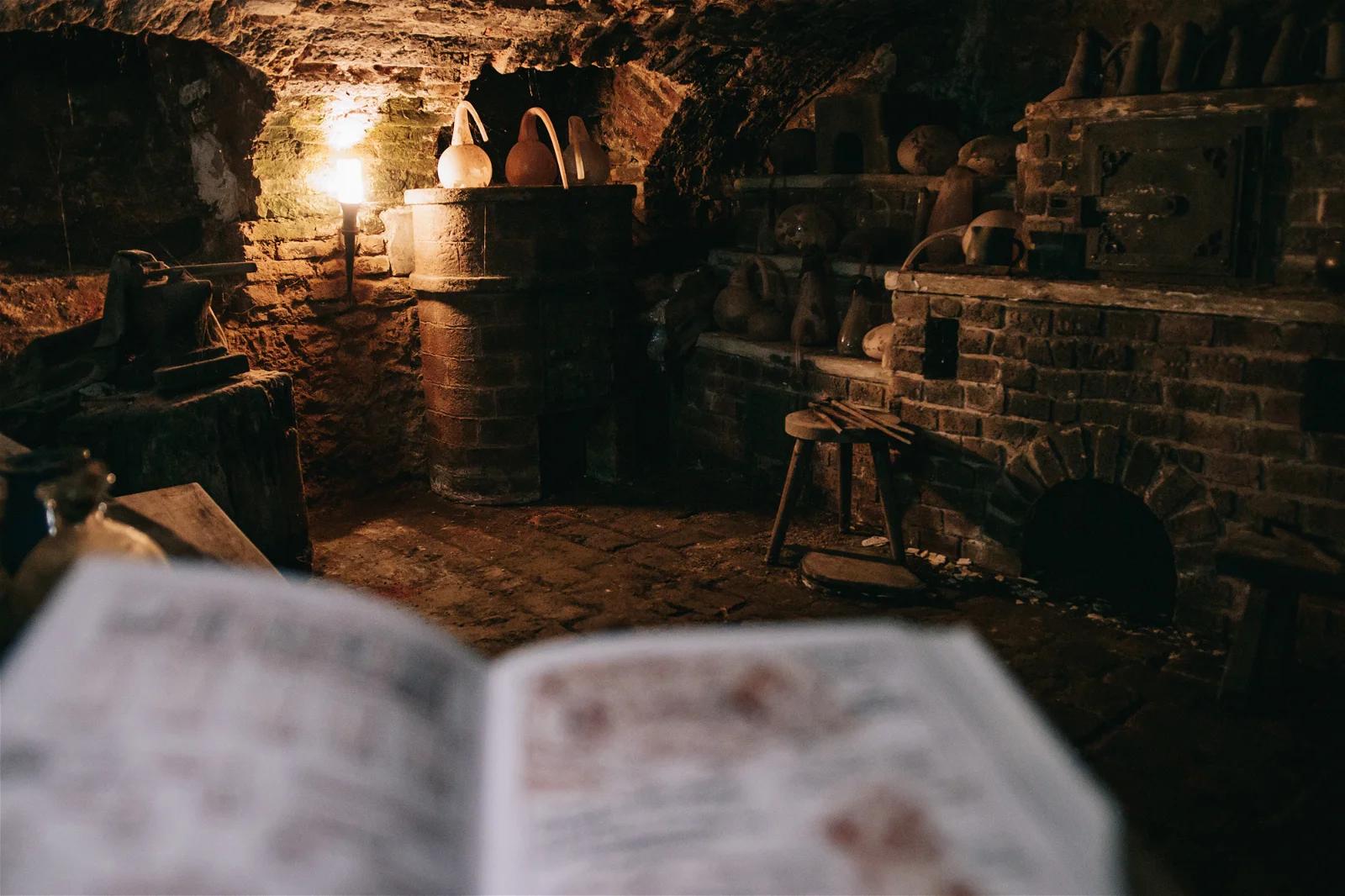 A close-up view of an open book with illustrations and notes, in a blurred, candlelit background suggesting a historical setting.
