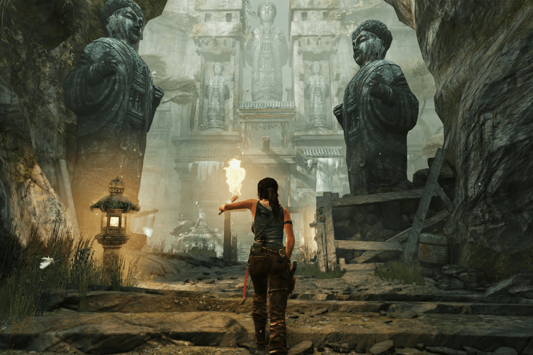 Lara Croft walks through an ancient temple courtyard, holding a torch as towering stone statues loom ahead.