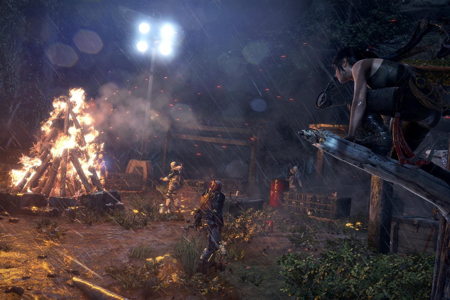 A nighttime scene from a Tomb Raider game capturing Lara Croft stealthily preparing to ambush an enemy by a bonfire in a rain-soaked, makeshift camp.