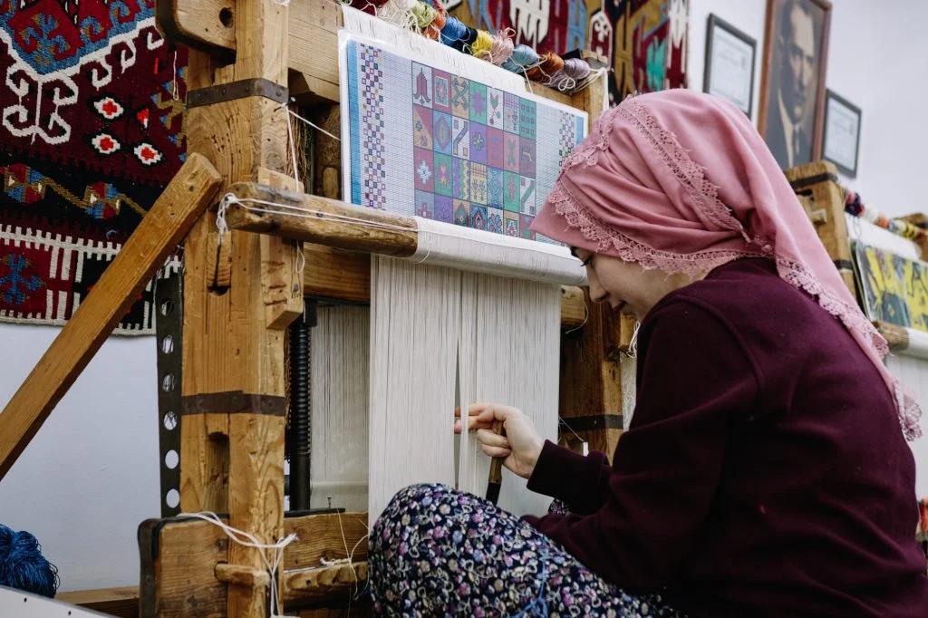 A woman in traditional attire is weaving on a loom, with intricate designs and textiles surrounding her.