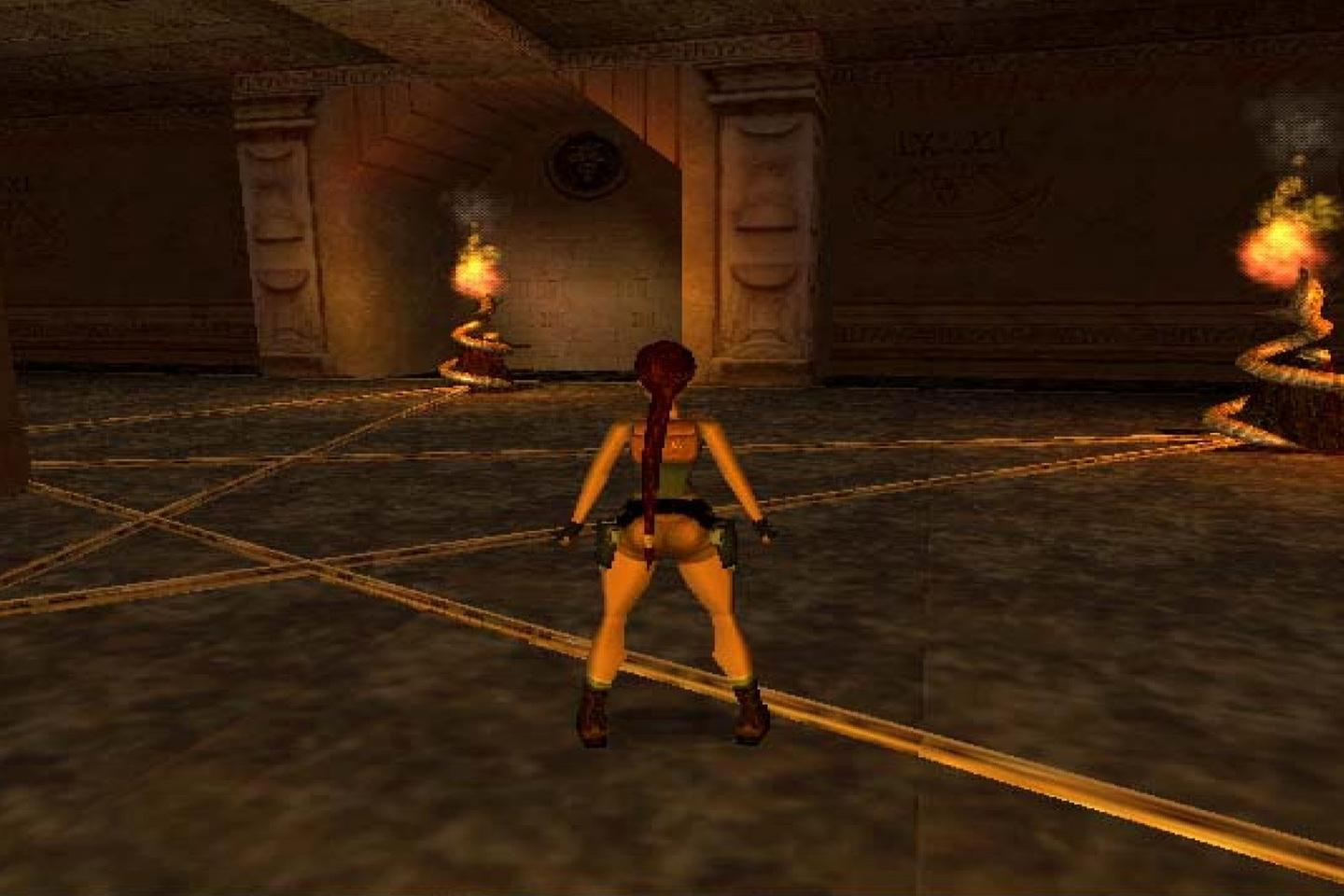 An in-game image of Lara with red hair and a yellow-black outfit standing in a dimly lit room with torches on the walls and hieroglyphic inscriptions.