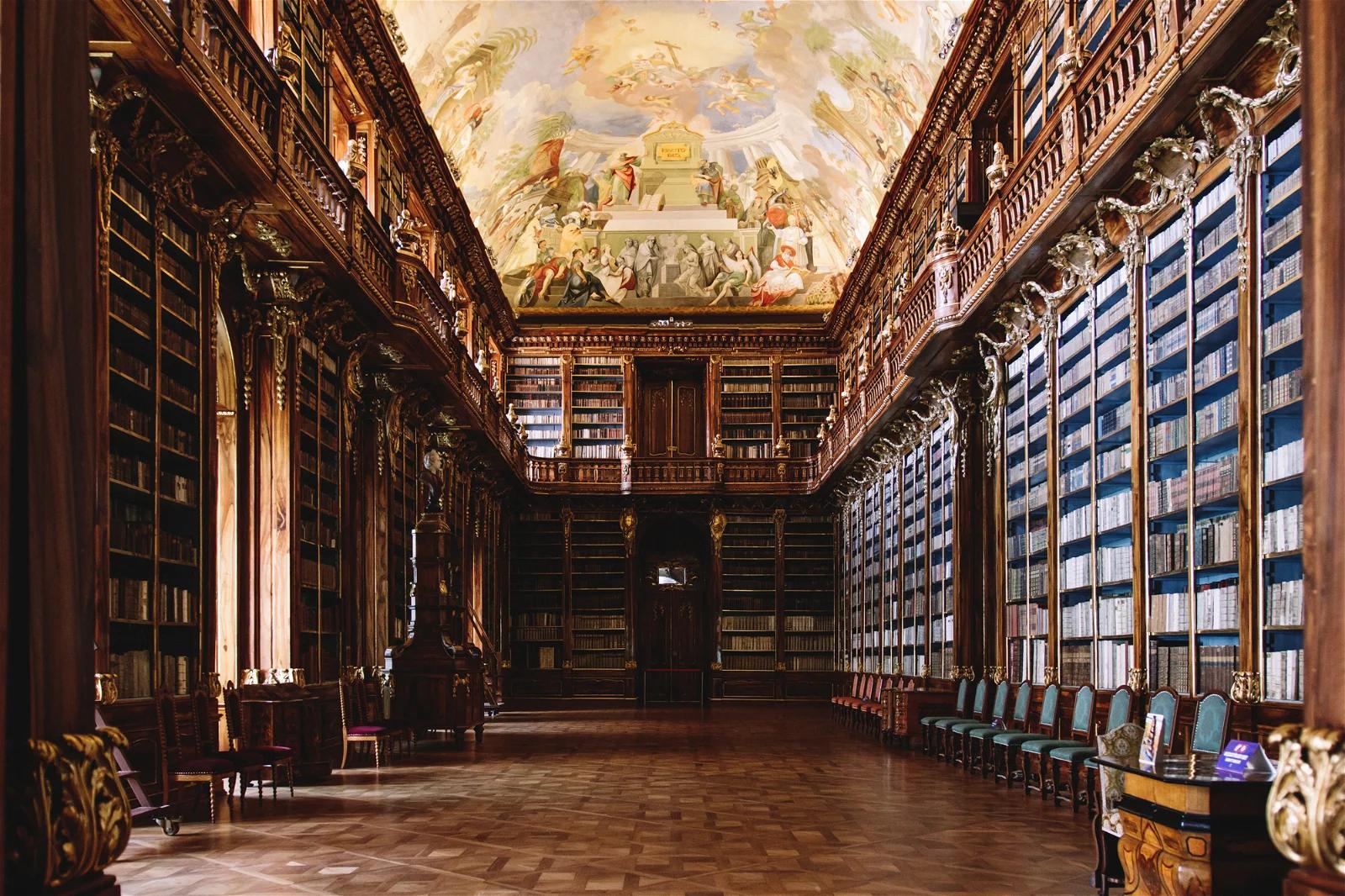 An elegant old library room with a high, painted ceiling and wall-to-wall bookshelves filled with blue-bound books, reflecting a classical European architecture.