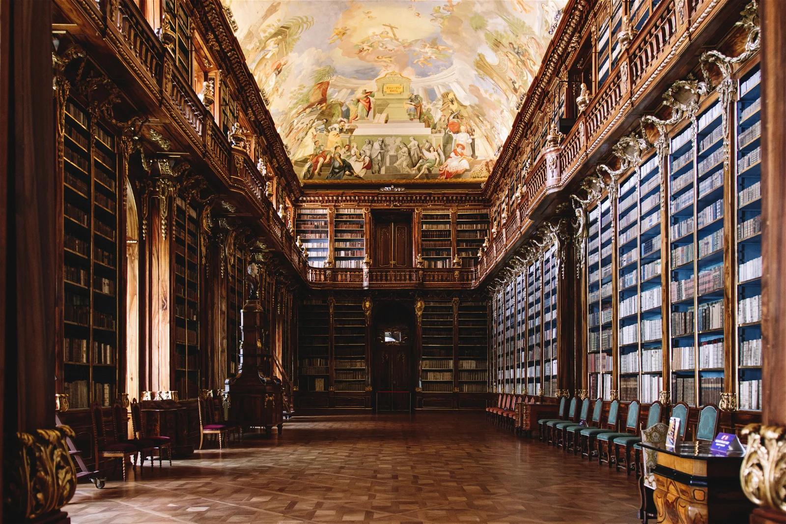 An elegant old library room with a high, painted ceiling and wall-to-wall bookshelves filled with blue-bound books, reflecting a classical European architecture.

