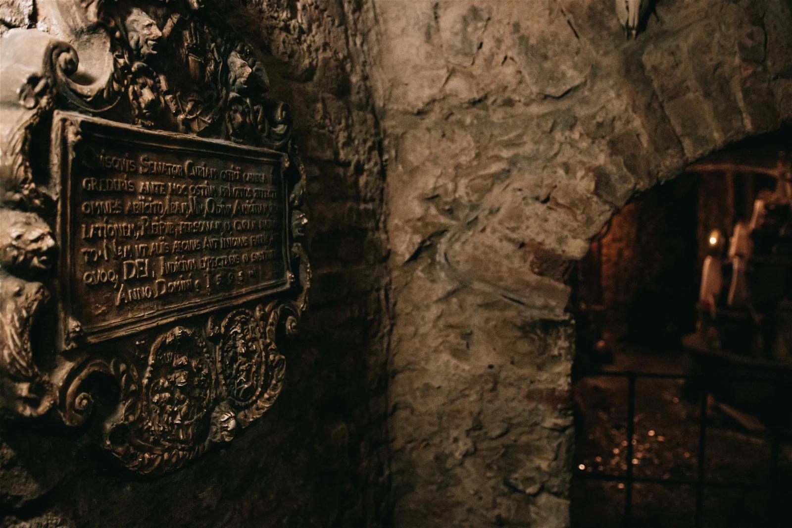 A plaque with inscriptions mounted on a stone wall in a dimly lit, historic-looking interior space.

