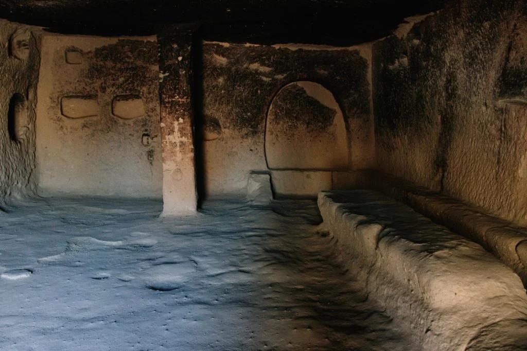 An underground room carved into the rock with niches and an arched doorway, resembling ancient living quarters.